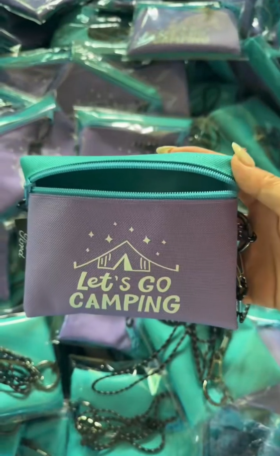 Custom purple cosmetic pouch with teal zipper and "Let's Go CAMPING" print, shown during bulk packaging at Bincai factory.